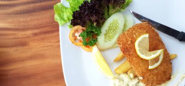 Kızarmış Dolly Fish bifteği taze organik sebze, yeşil lahana, dilimlenmiş salatalık, domates ve limon, kızarmış patates, krema sosu ve bıçak ahşap masada beyaz tabakta, sol kopya alanı ile birlikte.. 