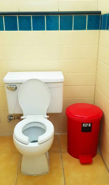 Flush toilet with red trash can on yellow tile in bathroom, restroom. Public washroom and clean room. Design and ceramic object