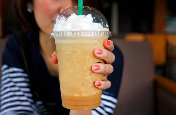 Asian woman in blue casual t-shirt holding a plastic glass of cold espresso Frappuccino with whipped cream on top and her orange nails. Starbucks is premium coffee. Blended drinking and refreshment