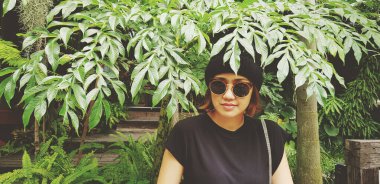 Asian woman is smiling, wearing sunglass and wool hat sitting on wooden bench under tree with green plant background. Girl in casual dress style in vintage tone color with copy space.	