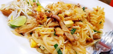 Tayland usulü kızarmış erişteyi ya da Tayland yemeğini kalamar, ahtapot ya da mürekkep balığı dilimlenmiş limon, sebze, tofu ve beyaz tabakta paslanmaz çelik kaşıkla kapat. Tayland 'da ünlü Asya yemekleri. 