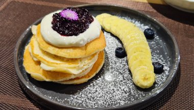 Krema ve yaban mersini reçeli ile kaplanmış krep, muzla servis edilmiş ve siyah tabakta krema şekeri ile tozlanmış. Gri tabakta meyveli çöl.