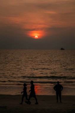 Dikey fotoğraf dramatik kırmızı gün batımı, sisli bulutların arkasında güneş yürüyen erkek ve kadın siluetleri, deniz kenarında duran bir adam..
