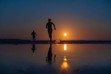 Gün batımında plajda koşan iki kişi. Güneş ufukta batarken yansımaları durgun sulara yansıyor. Su, koşan insanlara sıçrıyor.