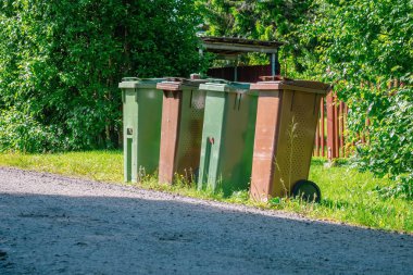 Güneşli bir yaz gününde dört çöp bidonu, temizlik işçileri tarafından boşaltılmayı bekliyor. Kutular plastikten yapılmış ve farklı renkte.