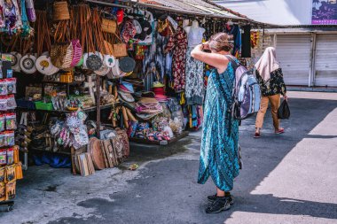 Mavi elbiseli ve sırt çantalı bir turist, Tanah tapınağı yakınlarındaki bir pazar yerinde renkli el sanatları çekerek canlı kültürü, 2020-01-05, Tanah Lot, Bali, Endonezya