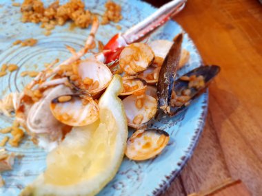 İstiridye, midye, Norveç ıstakozu, pirinç ve bir dilim limon, ahşap bir masadaki açık mavi tabakta duruyor. Lezzetli bir yemeğin kalıntılarını sergiliyorlar.