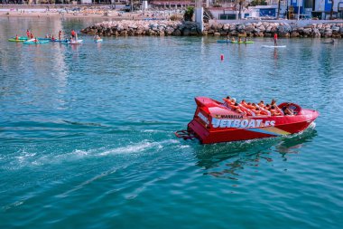 Canlı kırmızı sürat teknesi sakin mavi sularda palmiye ağaçları, modern binalar ve uzak dağlarla dolu manzaralı bir sahil şeridinde yarışıyor. PUERTO BANUS, Marbella 19 HAZİRAN 2022