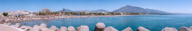 Yoğun bir kumsalın panoramik manzarası, sakin mavi deniz, kıyı binaları, ve berrak bir gökyüzünün altında görkemli bir dağ.