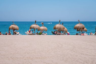 İnsanlar kumsalda güneşli bir günün tadını çıkarıyorlar saman şemsiyelerin altında, berrak mavi denizin kenarındaki sandalyelere uzanmış uzaktaki bir tekneyle.