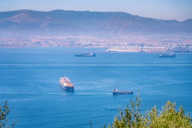 Kargo gemileri Cebelitarık körfezinde, Cebelitarık körfezinin karşısında yer alan büyük bir liman şehrinde, güneşli bir günde, açık mavi sularda ve arka planda dağlarla yol alıyorlar.