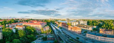 Gün batımında Stockholm 'ün şehir manzarasını yakalayan panoramik manzara, bir tren istasyonu, konut binaları, yemyeşil alanlar, ve arka planda biraz su, Stockholm, İsveç