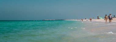 Dubai Kite sahilinde güneşli bir günün tadını çıkaran ziyaretçiler, turkuaz suyun kumlu kıyılarla buluştuğu kıyı şeridi boyunca yürüyorlar. Dubai BAE 27 Mart 2025