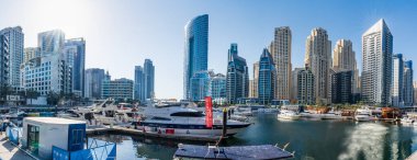 Dubai Marina 'nın panoramik manzarasında, parlak güneş ışığı altındaki modern gökdelenlerin zeminine kenetlenmiş lüks yatlar var. Dubai BAE 27 Mart 2025