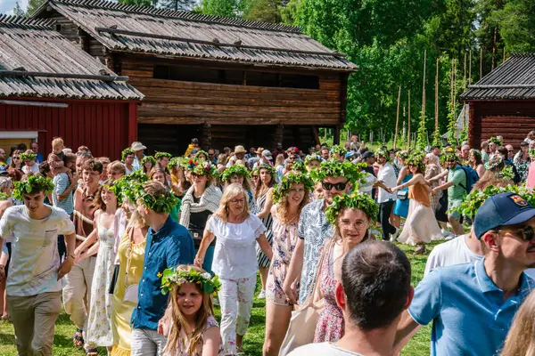 Turistler ve yerliler, Gammlia, Umea, İsveç 'te düzenlenen yaz kutlamaları sırasında çiçek çelenkleri giyerek ve festival atmosferinin tadını çıkararak bir zincirin içinde el ele tutuşuyorlar. UMEA, GAMLIA, İSVEÇ 23 HAZİRAN 2023