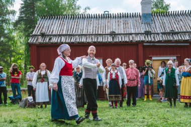 Gammlia, Umea 'da yaz ortası kutlamaları sırasında geleneksel İsveç kıyafetleriyle dans eden çift neşeli izleyiciler ve klasik kırmızı ahşap evle çevrili. UMEA, GAMLIA, İSVEÇ 23 HAZİRAN 2023
