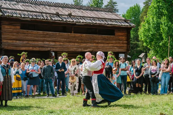 İsveç 'in Umea, Gammlia şehrinde düzenlenen bir yaz kutlaması sırasında çiçek çelengi takan turistler ve yerliler geleneksel kıyafetlerle dans eden çiftleri izliyorlar. UMEA, GAMLIA, İSVEÇ 23 HAZİRAN 2023