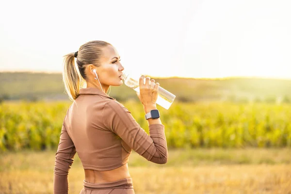 Young woman drinking water after jogging,healty and sport concept. Wears a beige sports suit and listens to music on headphones. High quality photo