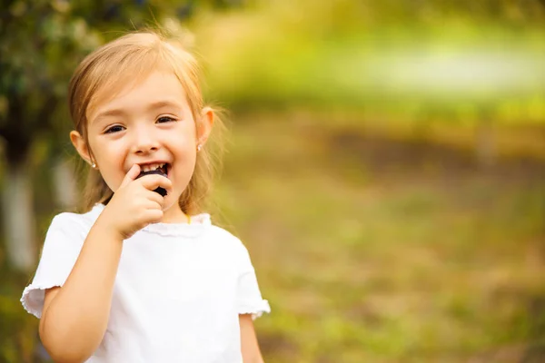 A girl bites a plum.A girl with a blonde hair the garden against a plum background.A toothless baby is trying to bite off a fruit.Funny and cheerful girl tries plum.Hairstyle after curls. High quality