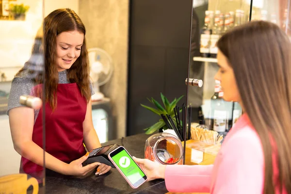 Female customer holding phone terminal make contactless mobile payment with smiling waitress barista saleswoman on coffeeshop counter, woman client pay in cafe with cellphone. High quality photo