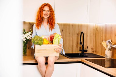 Çekici genç bir kadın mutfakta kollarında taze sebzelerle dikilirken çay içiyor. Yüksek kalite fotoğraf