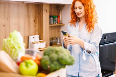 Genç mutlu kadın elinde beyaz bardakla mutfaktaki ahşap masada yeşil sebzelerin arasında otururken kameraya bakıyor. Yüksek kalite fotoğraf