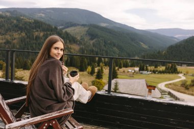Kadın, dağları görebileceğiniz ve renkli manzaranın tadını çıkarabileceğiniz kır evinin terasında bir fincan kahveyle tek başına oturuyor. Arka planda köknar ağaçları ve dağlar var.
