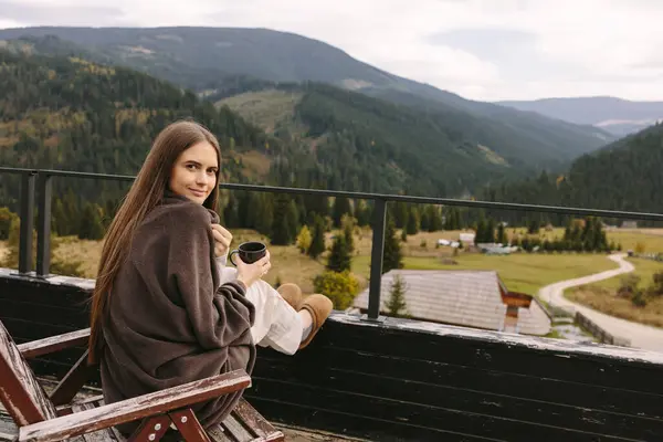 Kadın, dağları görebileceğiniz ve renkli manzaranın tadını çıkarabileceğiniz kır evinin terasında bir fincan kahveyle tek başına oturuyor. Arka planda köknar ağaçları ve dağlar var.