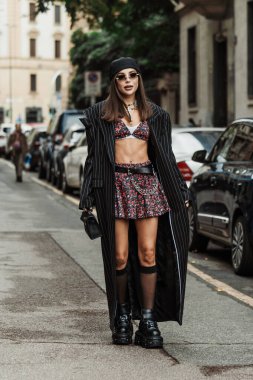 Karina Nigay siyah bir başlık, güneş gözlükleri, kolyeler, göbeğinde piercing, renkli bir çiçek desenli sütyen, aynı bel kıvrımlı mini etek, siyah bir Prada kemeri, ince beyaz çizgili siyah uzun bir palto, siyah deri bir çanta giyer., 