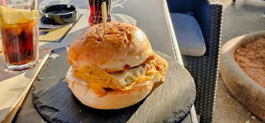 Ağzını sulandıran bir cheeseburger ve kıtır pastırma, siyah bir tabakta servis edilir, yanında da tazeleyici bir soda. Açık hava yemek ortamında yakalanmış. Sıradan bir yemek deneyimi için mükemmel..