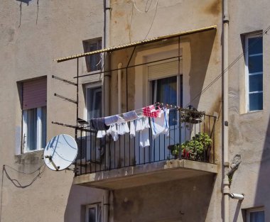 Konut balkonunda, bitkiler ve uydu anteninin yanında güneşte kurumuş asılı giysiler sergileniyor. Bu şehir manzarası bir şehir ortamında basitlik ve günlük hayatı yakalar..