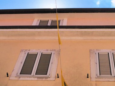 Canlı sarı bir bina cephesinde kepenkli pencereler ve çarpıcı mavi bir gökyüzü arka planı yer alıyor. Görüntü, minimalist mimari tarzda ve sakin bir atmosferle güneşli bir gün yakalıyor..