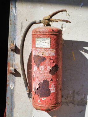 Eski ve yıpranmış boyalı bir yangın söndürücü gri beton bir duvara asılır. Güneş ışığı, gösteriş yaşı, ihmal ve kırsal dokularla aydınlatılır..
