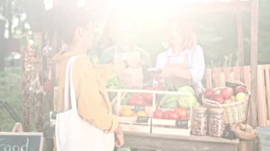 Genç bayan müşteri markette yiyecek satan çiftçi ailesinden market alışverişi yapıyor. Erkek ve kadın tezgahta ekolojik sebze satıyorlar. Müşteri kredi kartıyla ödeme yapıyor.
