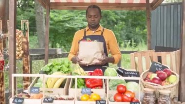 Yerel gıda pazarında organik sebze satan mutlu kıdemli çiftçi. Afro-Amerikalı bir adam kameraya gülümseyen sebzeler ve çiftlik ürünleriyle kağıt torba veriyor. İş konsepti