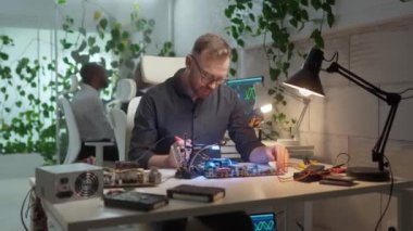 Modern ofiste bilgisayar lehimleyen çekici profesyonel teknisyen portresi. Hizmet merkezinde başarılı bir erkek çalışan mikroçipleri tamir ediyor gözlük takıyor ve bileşenler üzerinde çalışıyor..
