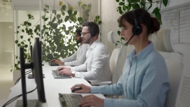 在呼叫中心工作的年轻专业人士的侧视图肖像 英俊而快乐的顾客支持人员 带着耳机坐在桌子边 与同事们一起使用电脑 微笑着看着相机 — 图库视频影像