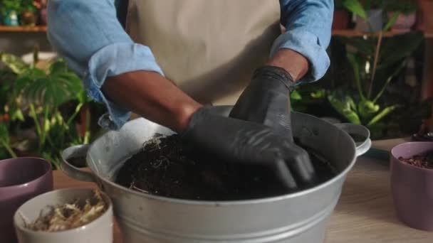 由快乐的职业非洲裔美籍男性花匠戴着手套和围裙微笑 将土壤与肥料混合在一起的特写镜头 迷人的非洲男人在花店工作 — 图库视频影像