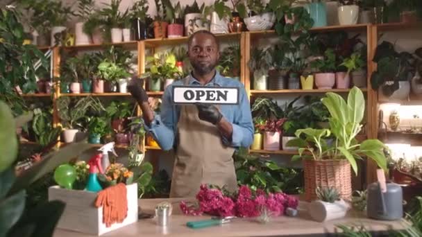 迷人而快乐的非洲年轻人站在花店边 当花店管理员 手持紧闭的标志 微笑着看着相机 漂亮快乐的职业男性 工作愉快 — 图库视频影像