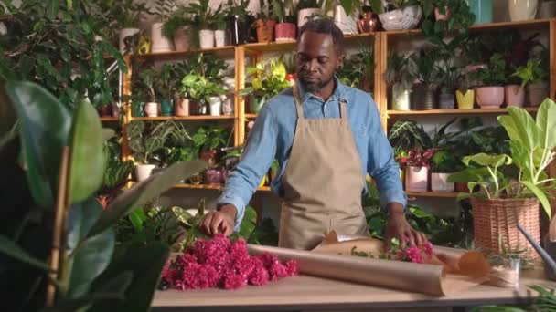 花屋の装飾と居心地の良い花店のカウンターにラッパーを使用して花の美しい花束を作るとして働く深刻な美しい大人のアフリカ人男性に焦点を当てました 花の概念 — ストック動画