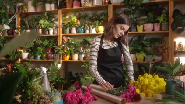 年轻而快乐的美丽女子在舒适的花店柜台上制作和装饰花束 迷人可爱的女花匠在智能手机上谈笑风生 — 图库视频影像