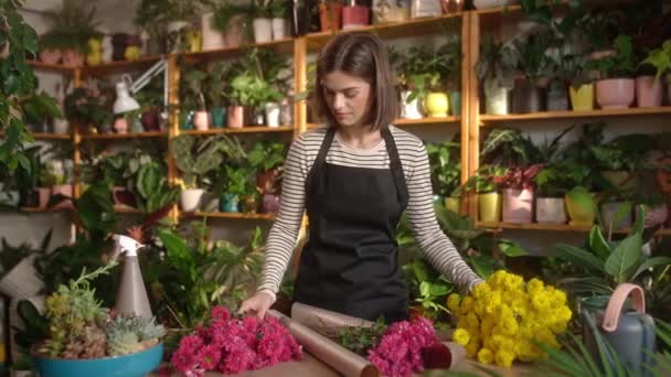 以专业迷人的年轻女性花设计师为焦点 在舒适的花店柜台上用包装纸装饰花束 漂亮的成年女性穿着围裙享受工作 — 图库视频影像
