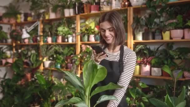 迷人的快乐快乐年轻的高加索女人在花店里用智能手机给植物拍照 她是个女花匠 美丽的成年女性微笑着工作愉快 — 图库视频影像