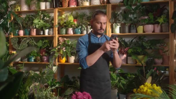 现代职业白人男子的画像 在花店用智能手机与顾客进行在线交流 漂亮的成年男工照顾花 — 图库视频影像