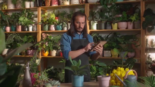 在花店工作的年轻貌美的成年人使用平板电脑在线监控植物 迷人的职业白种人男花匠发现了关于花的护理的信息 — 图库视频影像