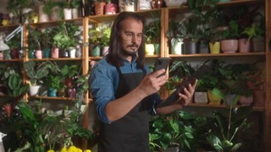 İnternette akıllı telefon kullanarak çiçekçileri denetleyen genç beyaz çiçekçi portresi. Önlüklü, güzel, uzun saçlı erkek işçi. Çiçek konsepti.
