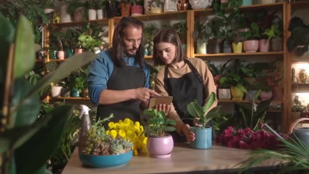 花の店で植物をオンラインで見てタブレットを使用して若いかわいいプロの女性花屋で働く大人のハンサムな長髪の男 エプロンをつけて２人の同僚が笑顔で — ストック動画