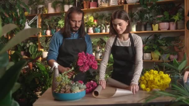 花屋のカウンターでラッパーを使用して花の花束を作る2つのプロの美しい若い花の装飾 かわいい女性とハンサムな男が一緒に働いています 花の概念 — ストック動画
