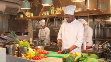 Kesme tahtasında bıçakla domates kesen konsantre Afro-Amerikan erkek aşçının portresi. Çekici Afrikalı şef arka planda meslektaşlarıyla lezzetli yemekler pişiriyor..