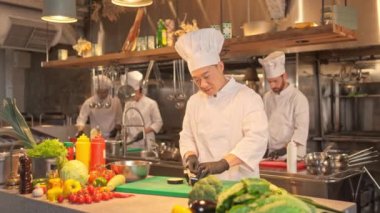 Odaklanmış yetenekli profesyonel Asyalı şef lezzetli salata pişiriyor bıçakla tahtayı keserken patlıcan doğruyor. Konsantre erkek aşçı. Mutfakta beyaz üniforma ve siyah eldiven giyiyor..